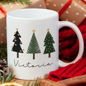 May include: White ceramic mug with a Christmas tree design. The mug features three stylized Christmas trees in black, green, and gold with gold stars on top. The name "Victoria" is written in black script below the trees.
