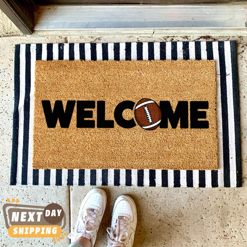 Pu&ograve; includere: Un zerbino rettangolare con la scritta "WELCOME" in lettere nere e una grafica di football americano. Il tappeto &egrave; beige con un bordo a righe bianche e nere. Le scarpe da ginnastica bianche sono visibili nella parte inferiore dell'immagine.