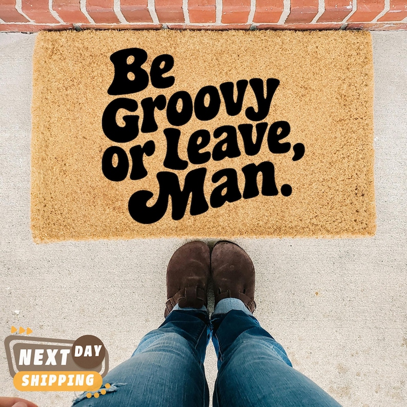 May include: A brown coir doormat with the text "Be Groovy or Leave, Man." in black lettering.