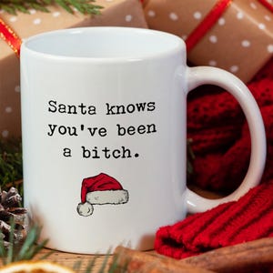 May include: White ceramic mug with the text "Santa knows you've been a bitch." in black, accompanied by a small Santa hat illustration. The mug has a curved handle and is set against a festive background with wrapped gifts and red knitwear.