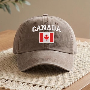Könnte beinhalten: Braune Baseballkappe mit dem Wort "CANADA" in Weiß auf der Vorderseite gestickt. Darunter befindet sich ein kleiner Aufnäher mit der kanadischen Flagge. Die Kappe ist aus einem weichen, abgenutzten Material.