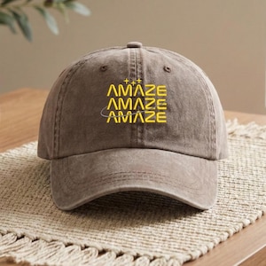 May include: A brown baseball cap with the word "AMAZE" embroidered in yellow. The word is stacked three times with a small star and a curved line design above and below the word. The cap has a curved brim and a fabric strap.