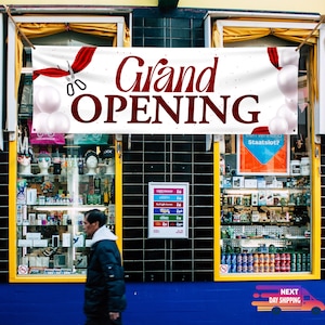 May include: A white banner with red ribbon and scissors announcing a grand opening. The banner is hanging above a shop front with a blue awning. The shop front has two large windows with merchandise displayed inside.