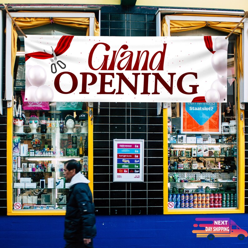 May include: A white banner with red ribbon and scissors announcing a grand opening. The banner is hanging above a shop front with a blue awning. The shop front has two large windows with merchandise displayed inside.