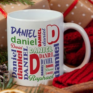 May include: White ceramic mug with a colorful design featuring the name "Daniel" repeated in various sizes and colors. The name "Raphael" is also included in the design.
