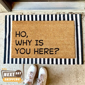 Puede incluir: Una alfombra de coco marr&oacute;n con ribete a rayas blancas y negras. La alfombra tiene el texto "HO, WHY IS YOU HERE?" impreso en negro.