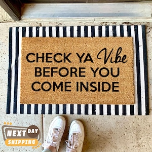 May include: A rectangular doormat with the text "CHECK YA VIBE BEFORE YOU COME INSIDE" in black. The mat has a natural brown center and a black and white striped border. White sneakers are visible at the bottom of the image.