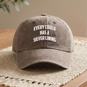 May include: A brown baseball cap with the phrase "EVERY CLOUD HAS A SILVER LINING" in white block letters. The cap is made of a soft, textured fabric and has a curved brim. The hat is sitting on a woven surface.
