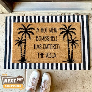 May include: A rectangular tan coir doormat layered over a black and white striped mat. The doormat features black silhouettes of four palm trees and the text A HOT NEW BOMBSHELL HAS ENTERED THE VILLA printed in a black sans-serif font across the center.