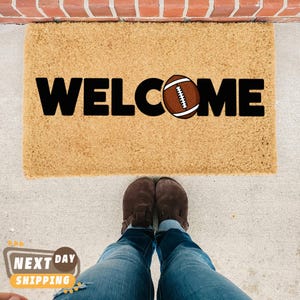 Pu&ograve; includere: Un zerbino rettangolare con la scritta "WELCOME" in grandi lettere nere. La "O" &egrave; sostituita da un pallone da football marrone e bianco. Il tappeto &egrave; realizzato in un materiale strutturato marrone chiaro.