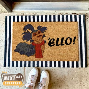 May include: A brown coir doormat with black and white stripes around the edges. The doormat features a cartoon illustration of a gray and blue creature with a red scarf and the text 'ello!'.