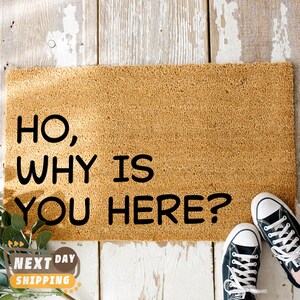 Puede incluir: Una alfombra de bienvenida marr&oacute;n con el texto "Ho, why is you here?" en letras negras. Dos zapatillas negras est&aacute;n en el suelo de madera delante de la alfombra.