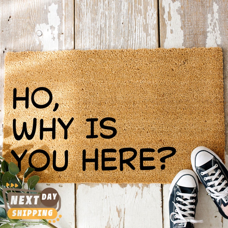 Puede incluir: Una alfombra de bienvenida marr&oacute;n con el texto "Ho, why is you here?" en letras negras. Dos zapatillas negras est&aacute;n en el suelo de madera delante de la alfombra.