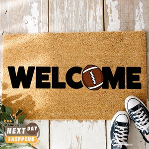 Pu&ograve; includere: Un zerbino rettangolare con la scritta "WELCOME" in grandi lettere nere. La "O" &egrave; sostituita da una grafica di pallone da football americano marrone. Il tappeto &egrave; beige e poggia su una superficie di legno bianco. Sono visibili delle scarpe da ginnastica nere.