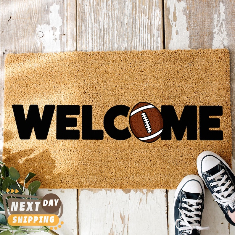 Pu&ograve; includere: Un zerbino rettangolare con la scritta "WELCOME" in grandi lettere nere. La "O" &egrave; sostituita da una grafica di pallone da football americano marrone. Il tappeto &egrave; beige e poggia su una superficie di legno bianco. Sono visibili delle scarpe da ginnastica nere.