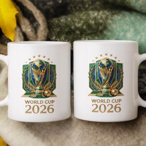 May include: Two white ceramic mugs featuring a graphic of the World Cup 2026 trophy. The design includes a gold and green geometric emblem with a laurel wreath, topped with stars. The text "WORLD CUP 2026" is printed in gold below the emblem.