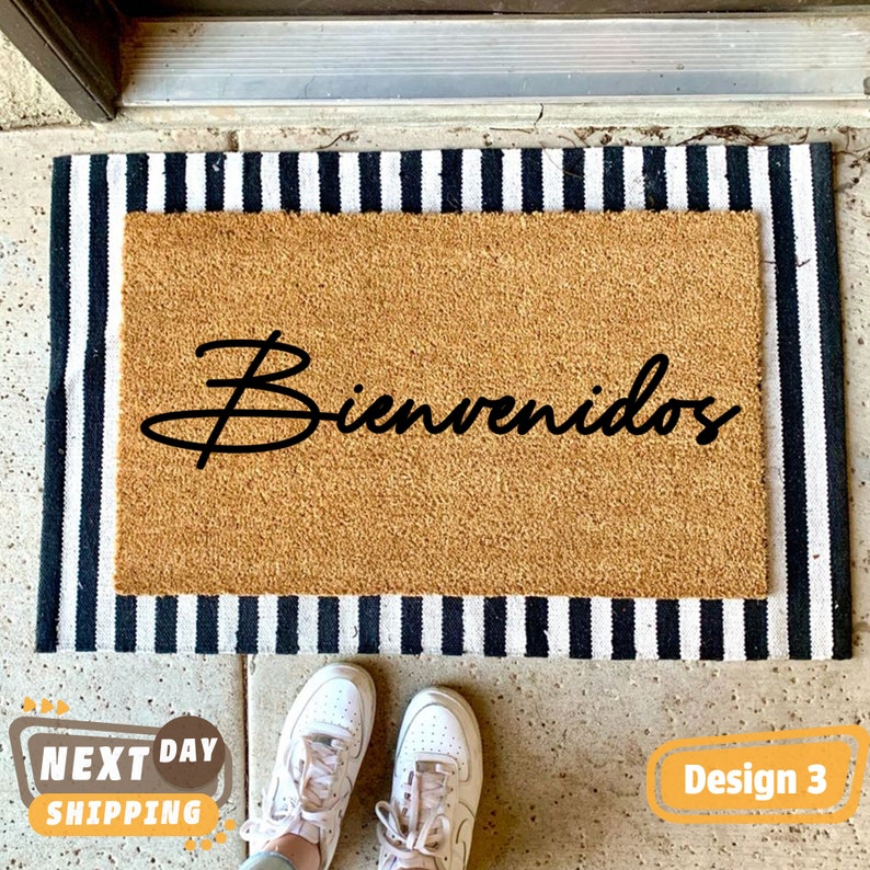 May include: A brown coir doormat with black and white striped edging. The doormat has the word "Bienvenidos" written in black cursive script.