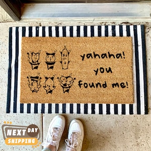 May include: A brown coir doormat with black and white stripes around the edges. The doormat has a black graphic of cartoon characters and the text "yahaha! you found me!"