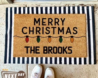 May include: A brown coir doormat with the text "Merry Christmas" and a string of colorful lights. The text "The Brooks" is below the lights.