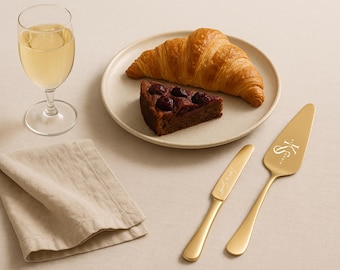 May include: A still-life image featuring a glass of white wine, a plate with a croissant and a slice of cake, a folded linen napkin, and gold serving utensils. The cake server is engraved with initials.