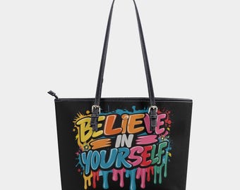 Op de afbeelding: Zwarte draagtas met lange handvatten en de tekst "BELIEVE IN YOURSELF" in kleurrijke graffiti-letters. De tas heeft een rechthoekige vorm en een ritssluiting. De handvatten zijn zwart met zilveren gespen.