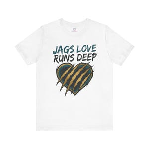 Puede incluir: Camiseta blanca con el texto "JAGS LOVE RUNS DEEP" en verde azulado y negro. La camiseta presenta un diseño de corazón con marcas de garras en dorado y verde azulado, creando un gráfico audaz.