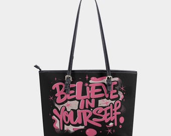 Op de afbeelding: Zwarte tote bag met roze opschrift "Believe in Yourself". Het opschrift is in graffiti-stijl met sterren en andere decoratieve elementen.