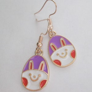 May include: A pair of gold-tone dangle earrings with purple and white enamel bunny rabbit egg charms. The bunny rabbit has a smiling face and red cheeks.