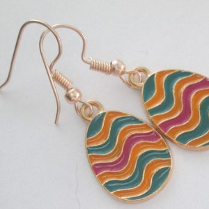 May include: A pair of gold-colored earrings with egg-shaped pendants. Each pendant features a colorful wavy design in teal, orange, and magenta. The earrings have a hook closure.