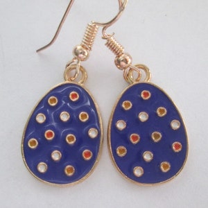 May include: A pair of dangle earrings featuring a blue egg-shaped design with gold trim. The eggs are decorated with white, orange, and gold dots. The earrings have gold-colored ear wires.