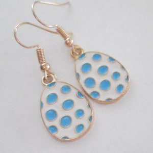 May include: A pair of gold-colored earrings with egg-shaped pendants. The pendants are white with blue polka dots and gold trim. The earrings have a hook closure.