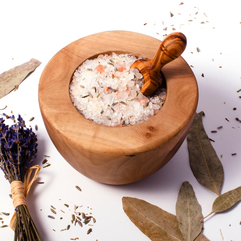 May include: A round wooden bowl filled with coarse pink Himalayan salt crystals mixed with dried lavender buds. A small wooden scoop rests inside the bowl. A bundle of dried lavender and several dried eucalyptus leaves are scattered nearby.