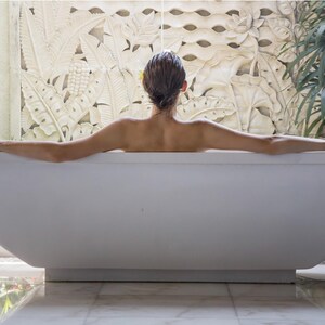 May include: A white rectangular freestanding bathtub with smooth, clean lines. The tub is positioned in front of a large, intricate stone wall carving featuring detailed botanical patterns of tropical leaves and flowers, creating a textured backdrop.