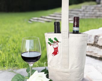 May include: A canvas wine tote with a Christmas stocking and holly design, holding two wine bottles. A glass of red wine and a white rose are in the foreground. The tote has a handle for easy carrying.