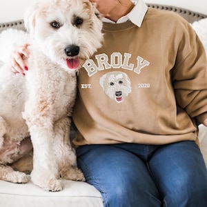 Puede incluir: Una persona lleva una sudadera de color marr&oacute;n claro con un dise&ntilde;o bordado en blanco que dice "BROLY EST. 2020" y presenta la cabeza de un perro. La persona est&aacute; sentada en un sof&aacute; blanco con un perro blanco.