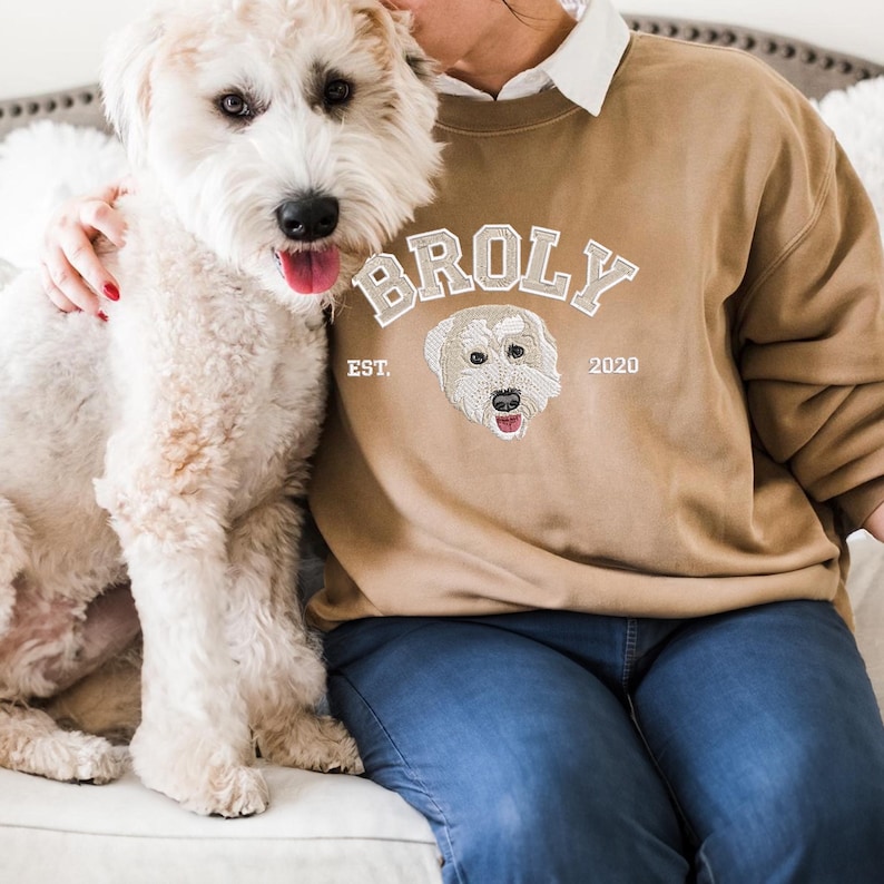 Puede incluir: Una persona lleva una sudadera de color marr&oacute;n claro con un dise&ntilde;o bordado en blanco que dice "BROLY EST. 2020" y presenta la cabeza de un perro. La persona est&aacute; sentada en un sof&aacute; blanco con un perro blanco.