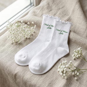May include: A pair of white ribbed crew socks featuring a ruffled top edge and green embroidered text that reads Maid of Honor Julia. The socks are made of a soft fabric and are designed with a classic fit, showcasing clean lines and embroidery.