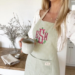May include: A green and white striped apron with a pink monogram and green floral accents. The apron has a pocket and a small tag that reads "Kitchen Cooking". The apron is worn with a white blouse and a beige strap.
