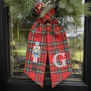 May include: A red plaid Christmas door decoration with a decorative bow and greenery. The decoration features a red plaid ribbon with an embroidered nutcracker and the letter "G". The ribbon is hanging on a glass door.