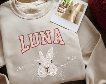 May include: A beige sweatshirt with a white rabbit embroidered on the front. The sweatshirt has the text "LUNA EST. 2022" embroidered on it.