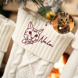 May include: White knitted Christmas stocking with a red embroidered cat illustration and the name "Nala" underneath. A photo of a tabby cat is in the background.