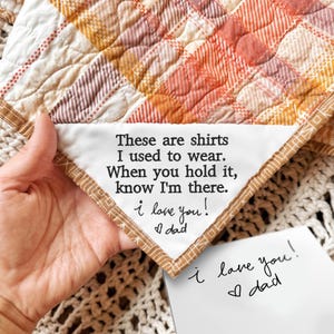 May include: A quilted blanket with a plaid pattern in shades of orange, yellow, and white. A white triangular patch with embroidered text reads "These are shirts I used to wear... I love you! Dad." A handwritten note says "I love you! Dad."