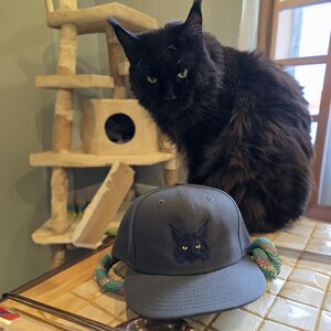 May include: A black cat with green eyes sits on a table next to a dark blue baseball cap with a black cat embroidered on the front.