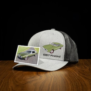May include: A gray trucker hat with a black mesh back and a green and white embroidered classic car design. The hat features the text "1967 Firebird" below the car graphic. A small photo of the car is also included.