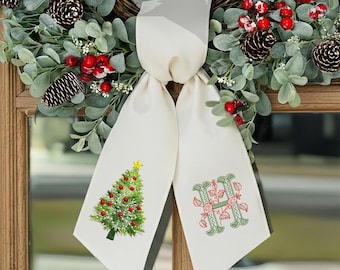 May include: A cream-colored Christmas bow with a green Christmas tree design on one side and the letter "H" on the other. The bow is hanging in front of a wreath with pinecones and red berries.