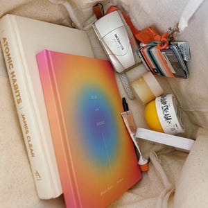 May include: A white tote bag filled with various items, including a book titled "Atomic Habits" by James Clear, a rainbow-colored journal with the words "Rise and Shine", a small silver camera, a striped pouch, a small jar of cream, and a yellow container with the words "Body Cream".