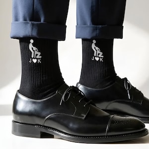 May include: A pair of black ribbed crew socks featuring a white graphic of two stick figures in a sexual position above the letters J and K separated by a heart symbol. The socks are worn with dark blue cuffed trousers and black leather dress shoes.