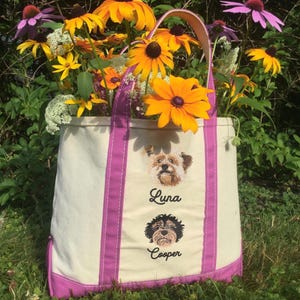May include: A white canvas tote bag with pink trim and two embroidered dog portraits. The top dog portrait is of a brown and white dog with the name "Luna" embroidered below. The bottom dog portrait is of a black dog with the name "Cooper" embroidered below.