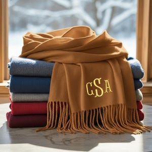 May include: A stack of folded blankets in various colors, topped with a tan scarf. The scarf has a fringe and gold embroidered initials. The blankets are blue, gray, red, and burgundy.