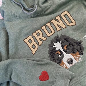 Puede incluir: Una sudadera con capucha verde con el nombre "BRUNO" bordado en beige en la parte delantera. Un perro de monta&ntilde;a bern&eacute;s est&aacute; bordado debajo del nombre. Un coraz&oacute;n rojo est&aacute; bordado en la manga izquierda.